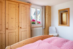 Une chambre avec une armoire en bois, une fenêtre donnant sur des maisons et des fleurs, un radiateur et une chaise blanche.