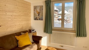 A room with a wooden wall, a brown sofa with a yellow pillow, a wooden coffee table, a window with green curtains, and a radiator. - Apartment 'Gästehaus Steiert Fewo Kuhbergblick' with Mountain View, Shared Garden and Wi-Fi (Lenzkirch)