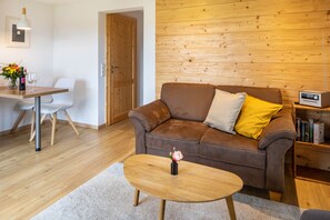 Un salon chaleureux avec un canapé marron, une petite table basse en bois, une table à manger blanche avec des chaises, et un mur en bois.