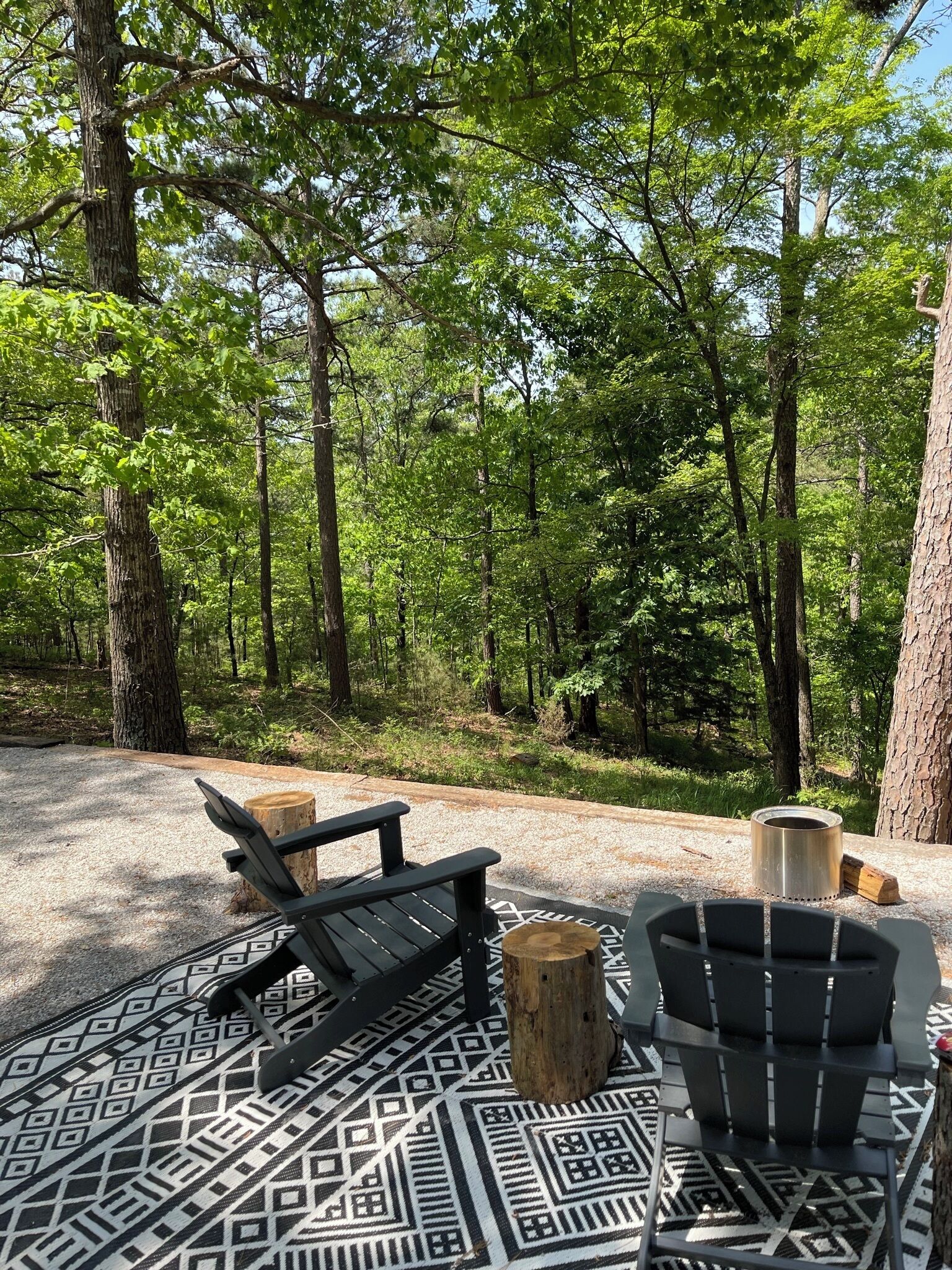 A forested area with outdoor seating including two Adirondack chairs and a bench, surrounded by trees and a patterned ground cover.