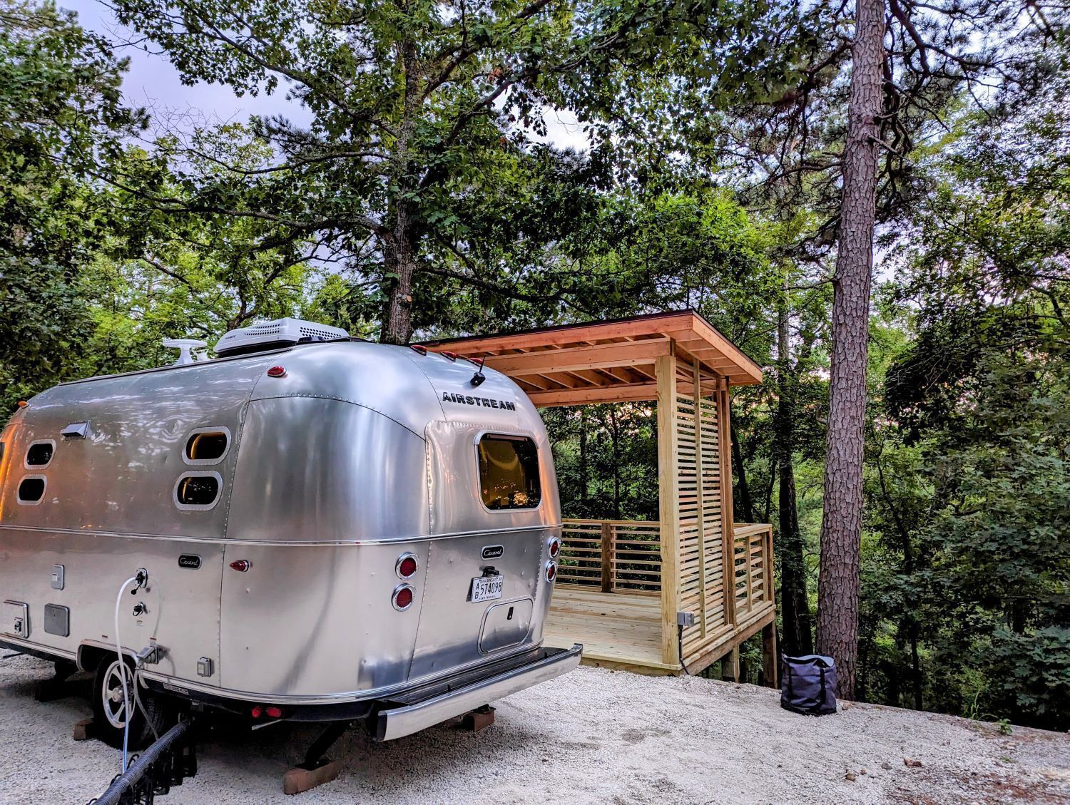 A silver Airstream trailer parked in a forested area with a wooden deck structure nearby.