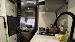 Une petite cuisine équipée d’un plan de travail, d’un évier et d’une cuisinière. On aperçoit, au fond, un lit avec une tête de lit et des oreillers.