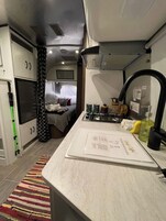 Una cocina compacta con una encimera, un fregadero y una estufa. Se ve una cama con cabecera y almohadas al fondo.