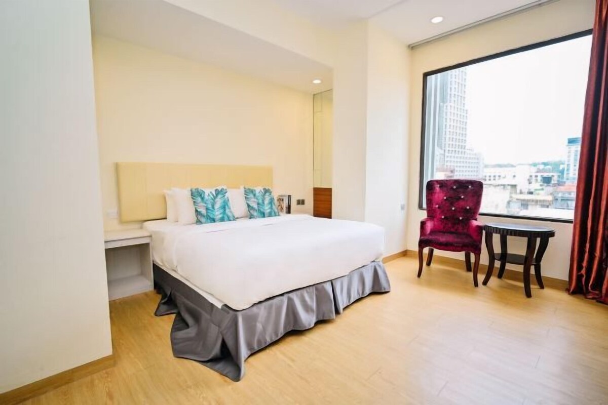 Habitación de hotel con una cama grande, un sillón rojo, una mesita y un ventanal con vistas a la ciudad.