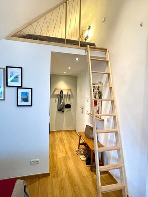 Ein Zimmer mit einer Holzleiter, einem Holztisch, einem Stuhl, einem Kleiderhaken und gerahmten Bildern an der Wand.