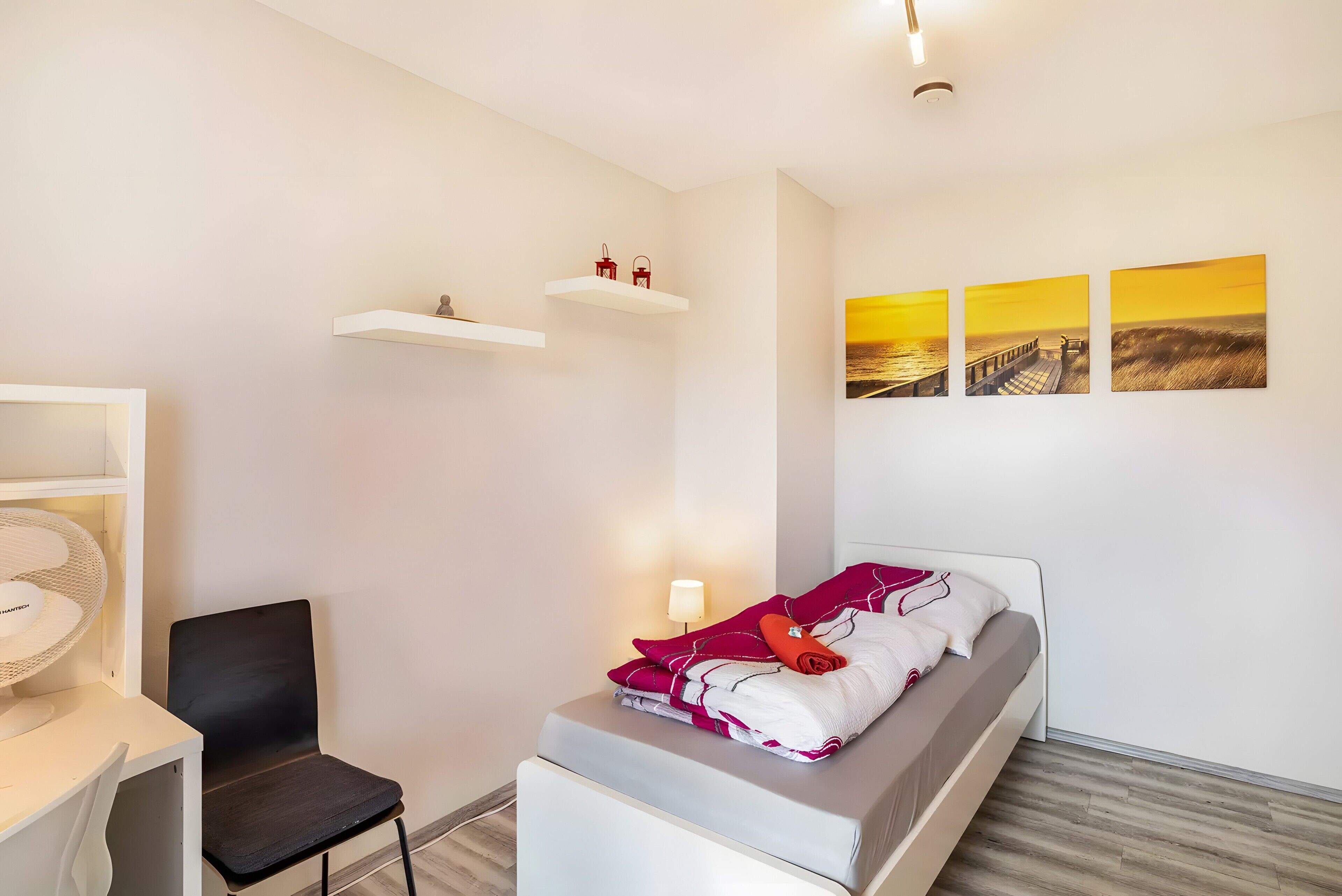 Een slaapkamer met een bed, een stoel, een bureau met lamp en wandplanken.