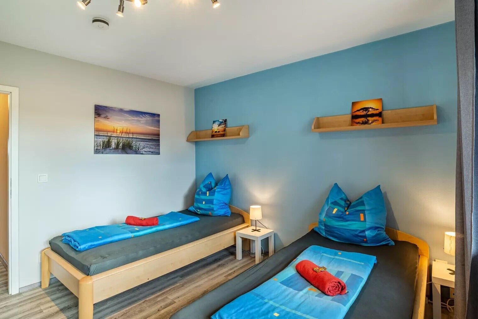A room with two single beds, a small bedside table with a lamp, a wall-mounted shelf with a book, and a painting of a sunset.