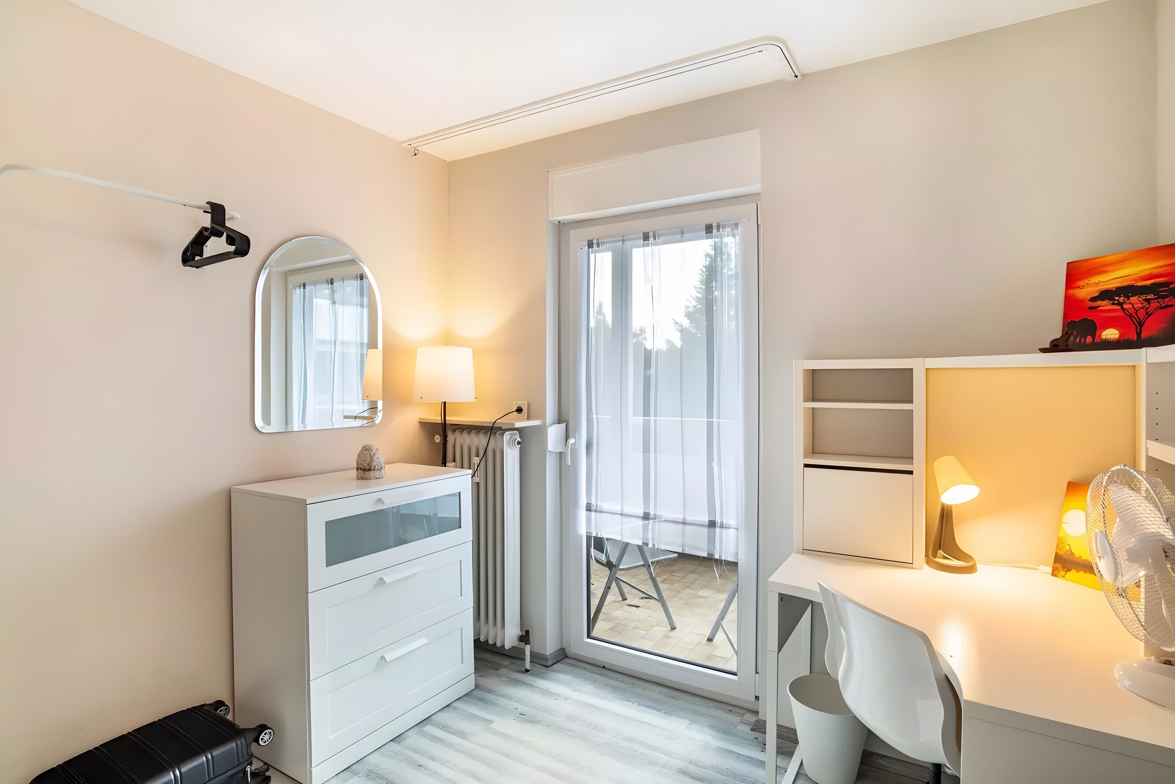 Un dormitorio moderno con un aparador blanco, un espejo, un escritorio con lámpara y un ventanal corredizo que da a un espacio exterior.