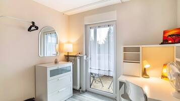 Un dormitorio moderno con un aparador blanco, un espejo, un escritorio con lámpara y un ventanal corredizo que da a un espacio exterior.