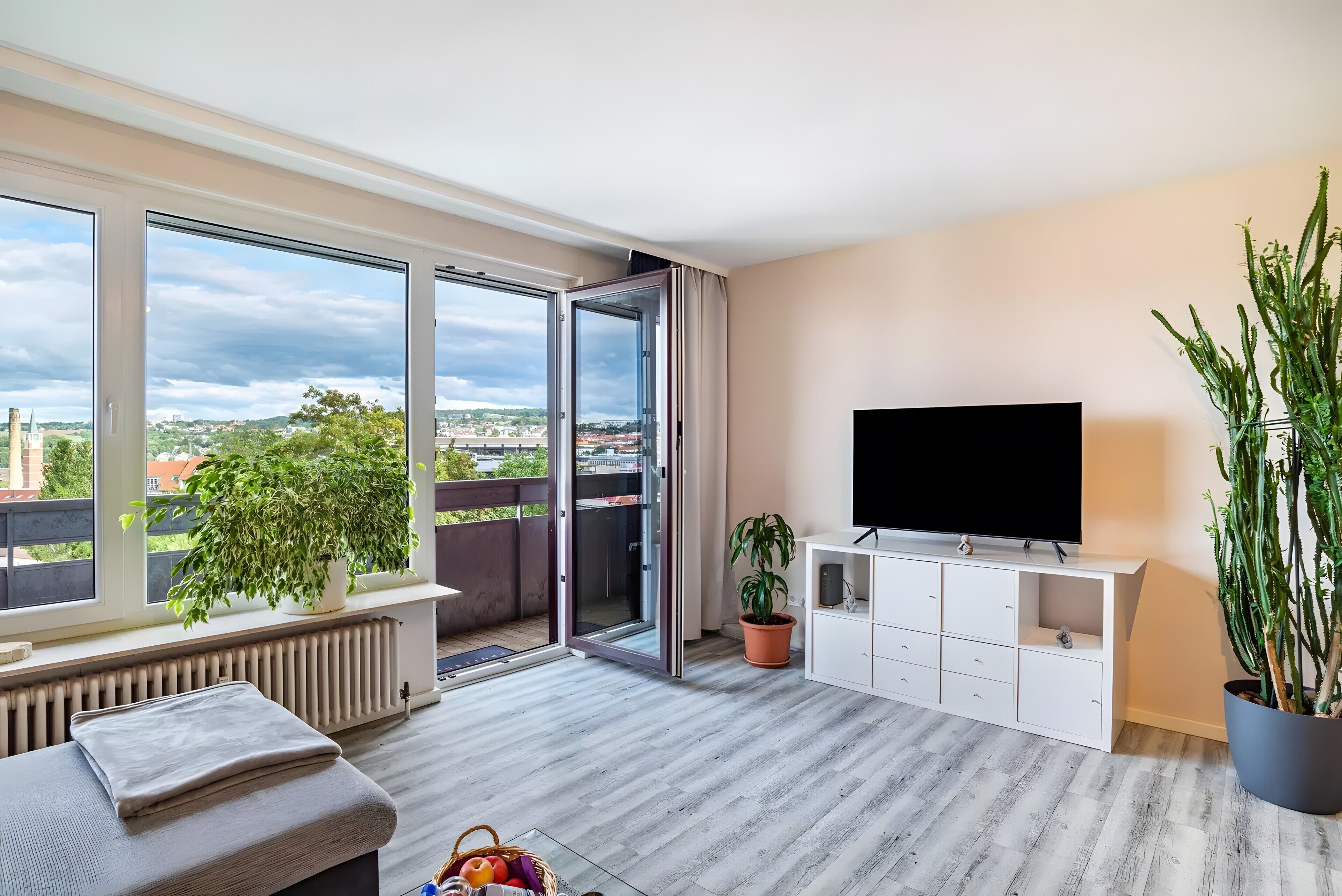 Een moderne woonkamer met een groot raam, een flatscreen televisie, een wit kastje en een potplant.