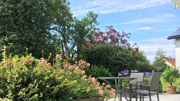 Een terras met een tafel en stoelen, omgeven door weelderige begroeiing en bloeiende planten.
