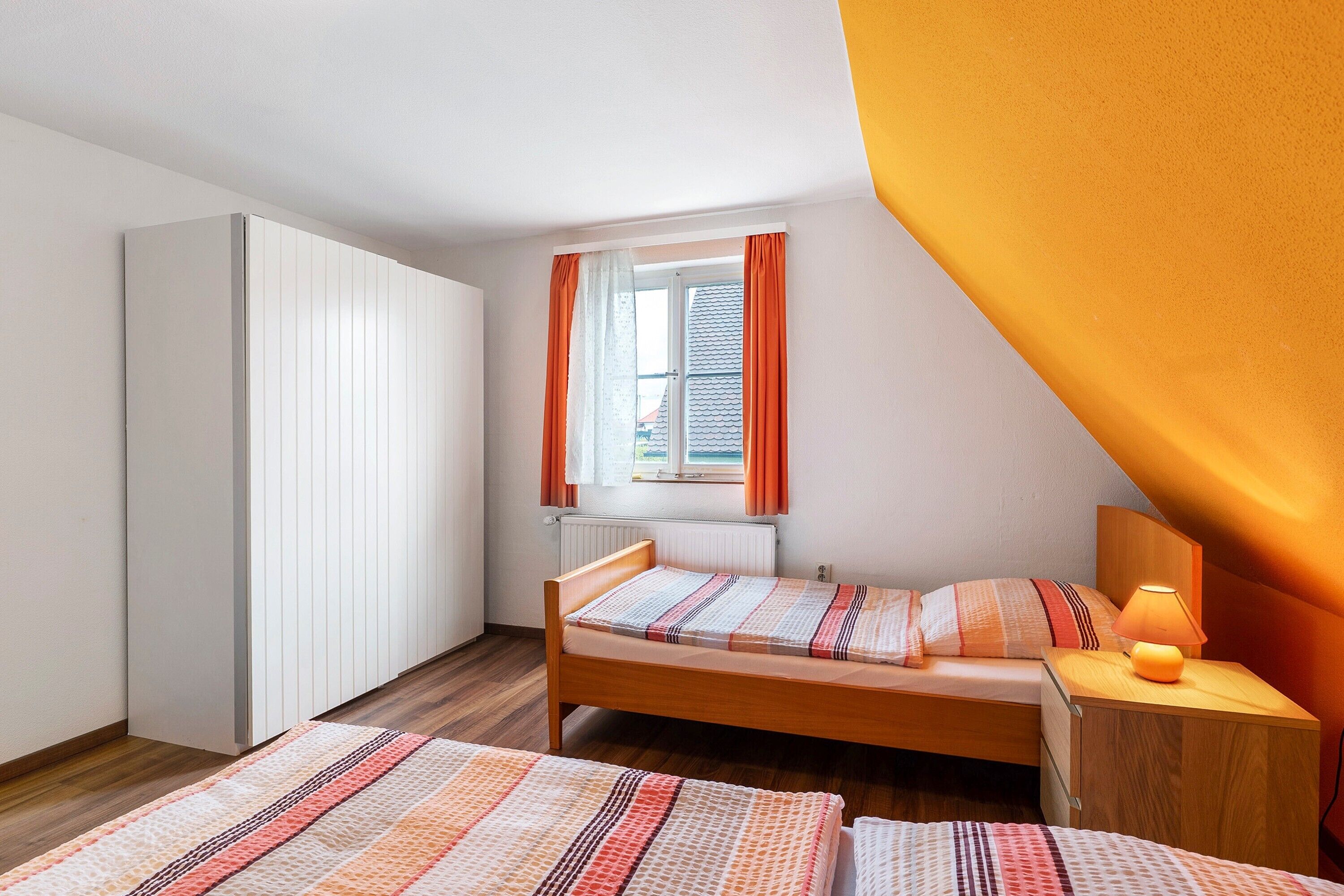 A bedroom with a bed, a nightstand, a lamp, a window with curtains, and a wardrobe.
