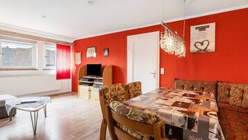 A room with a red wall, a dining table with patterned tablecloth, a sofa with a floral design, a wooden TV stand, and a chandelier.