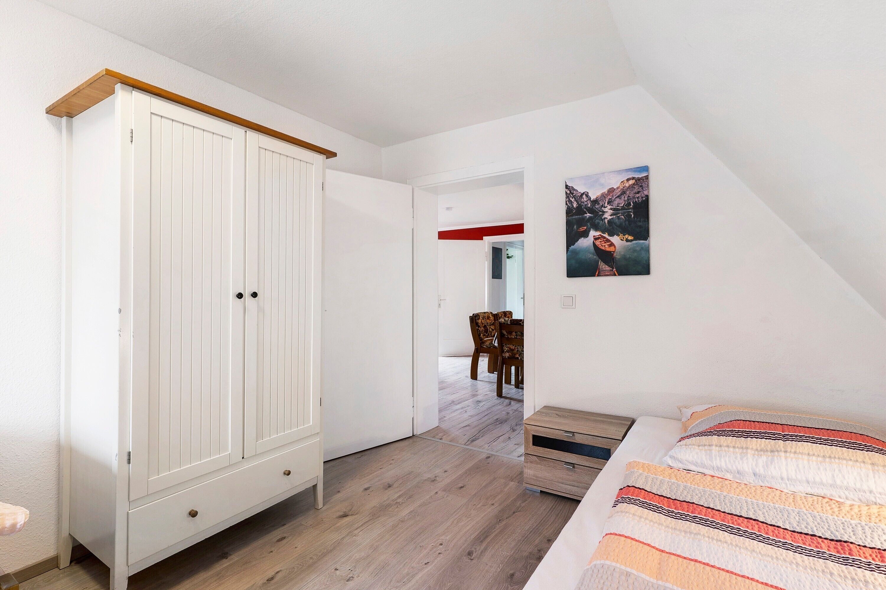 Ein Schlafzimmer mit weißem Kleiderschrank, einem Bett mit gestreifter Decke, Holzboden und einem Bild an der Wand.