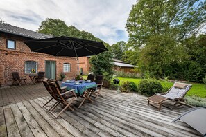 A wooden deck with a table and chairs, a large umbrella, and a brick building in the background.