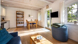 A spacious living area with a wooden ceiling, a dining table with chairs, a sofa, a TV mounted on the wall, and a window with curtains.