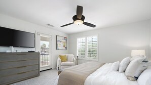 Une chambre à coucher comprenant un lit, une commode, un téléviseur, un ventilateur de plafond et des fenêtres équipées de stores.