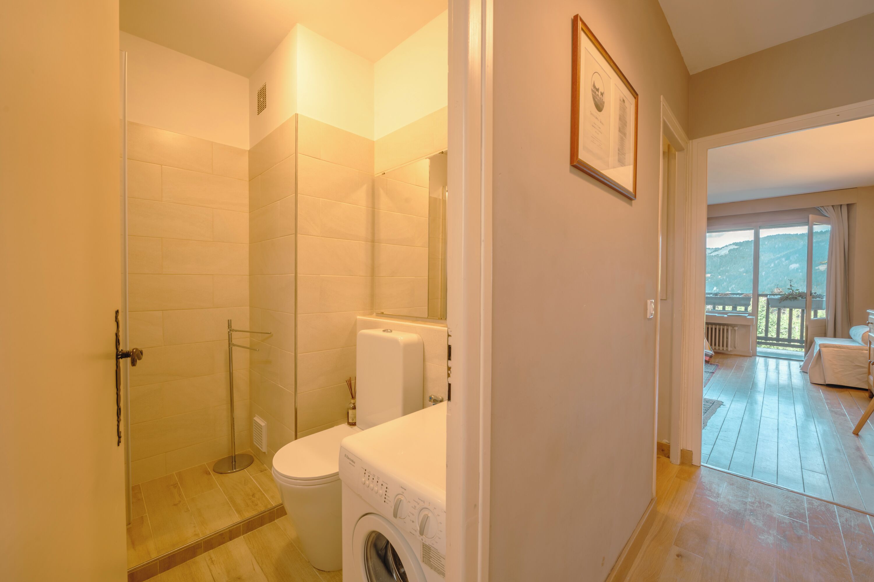 Un baño con ducha, inodoro y lavadora. Hay un cuadro en la pared y vistas a un balcón con terraza de madera.