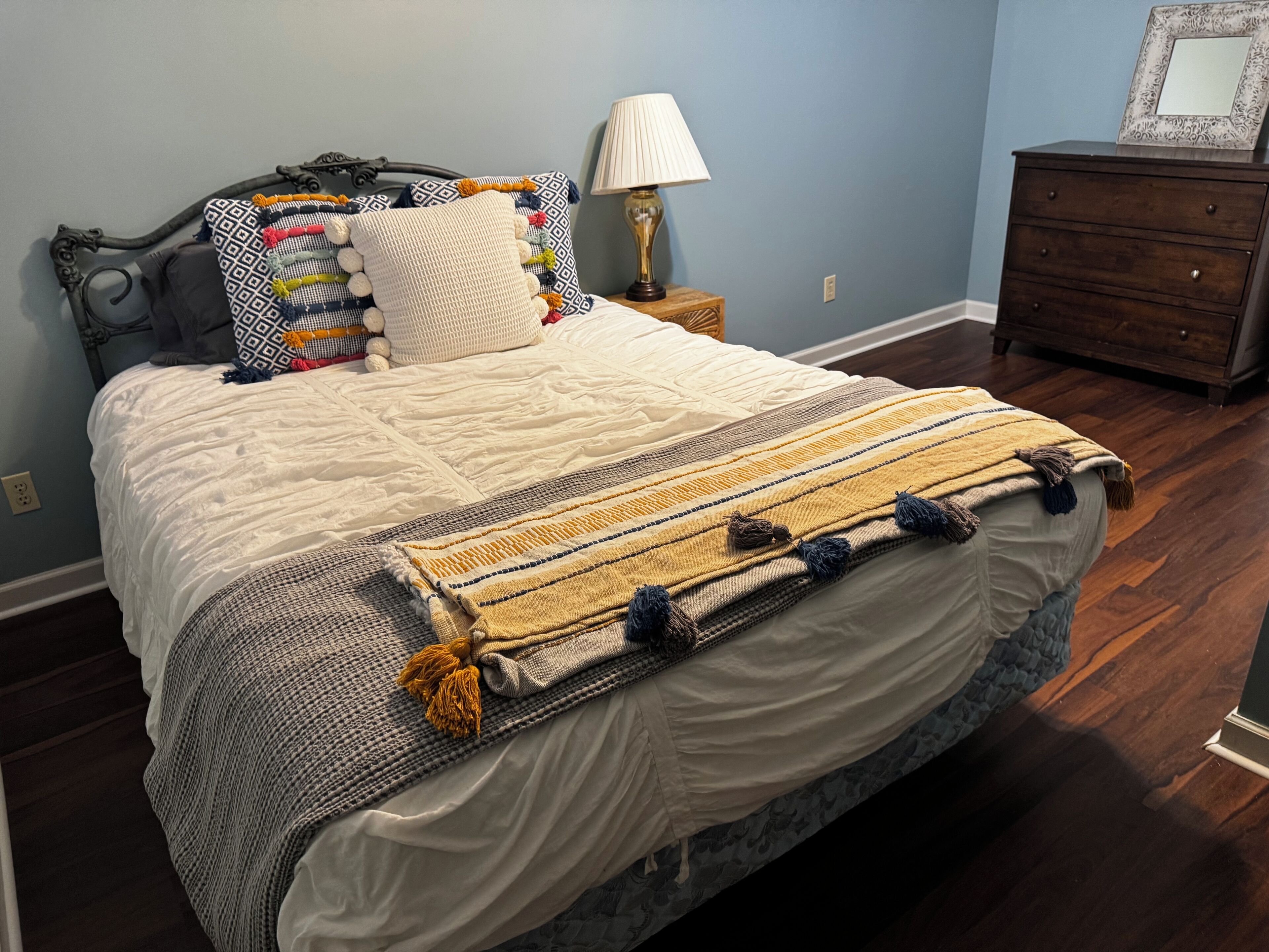 A neatly made bed with a patterned blanket and pillows, a bedside lamp, and a dresser against a blue wall.