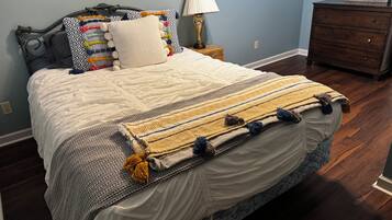 A neatly made bed with a patterned blanket and pillows, a bedside lamp, and a dresser against a blue wall.