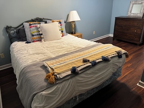 A neatly made bed with a patterned blanket and pillows, a bedside lamp, and a dresser against a blue wall.