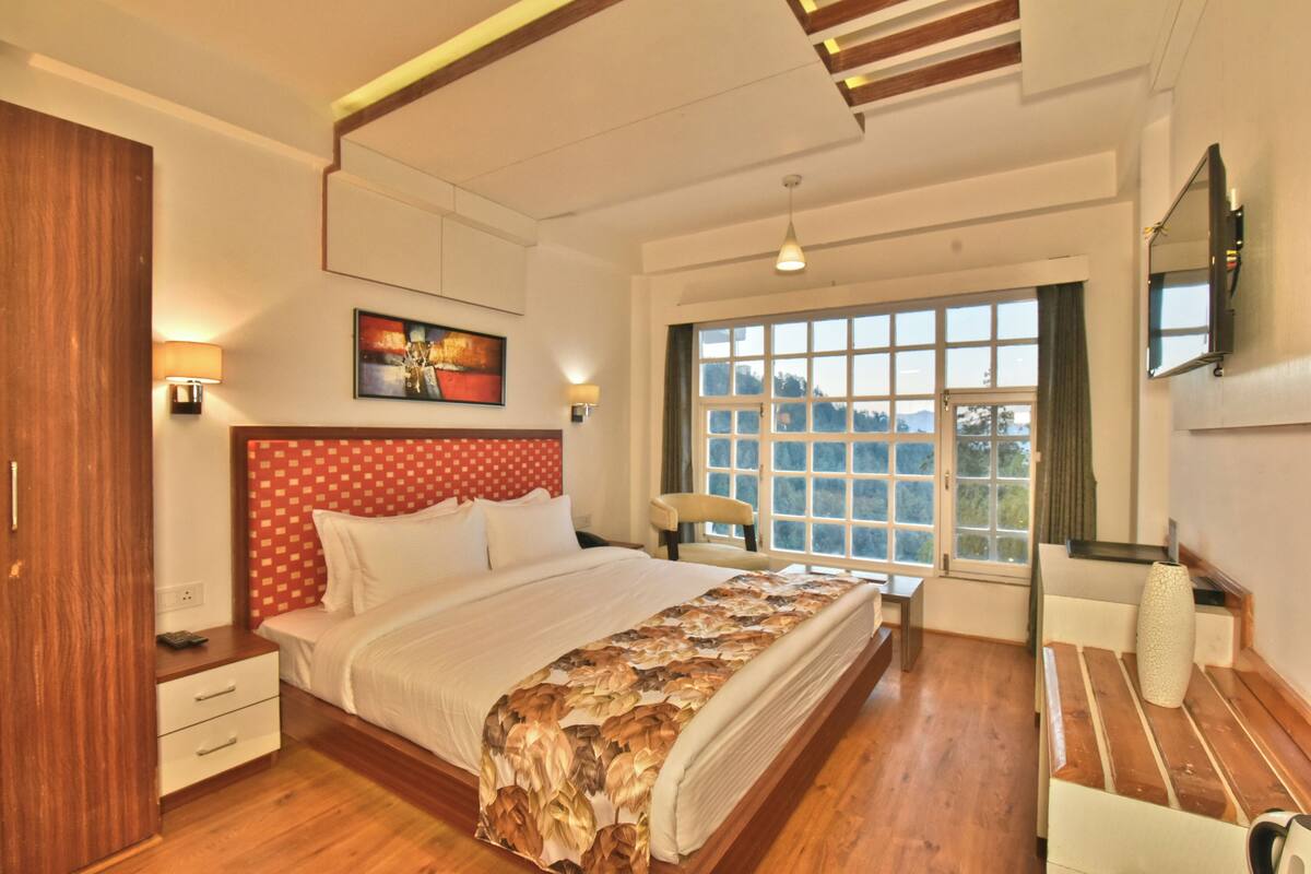A spacious bedroom with a large bed, wooden flooring, a red and white headboard, a window with a view, and a mounted TV.