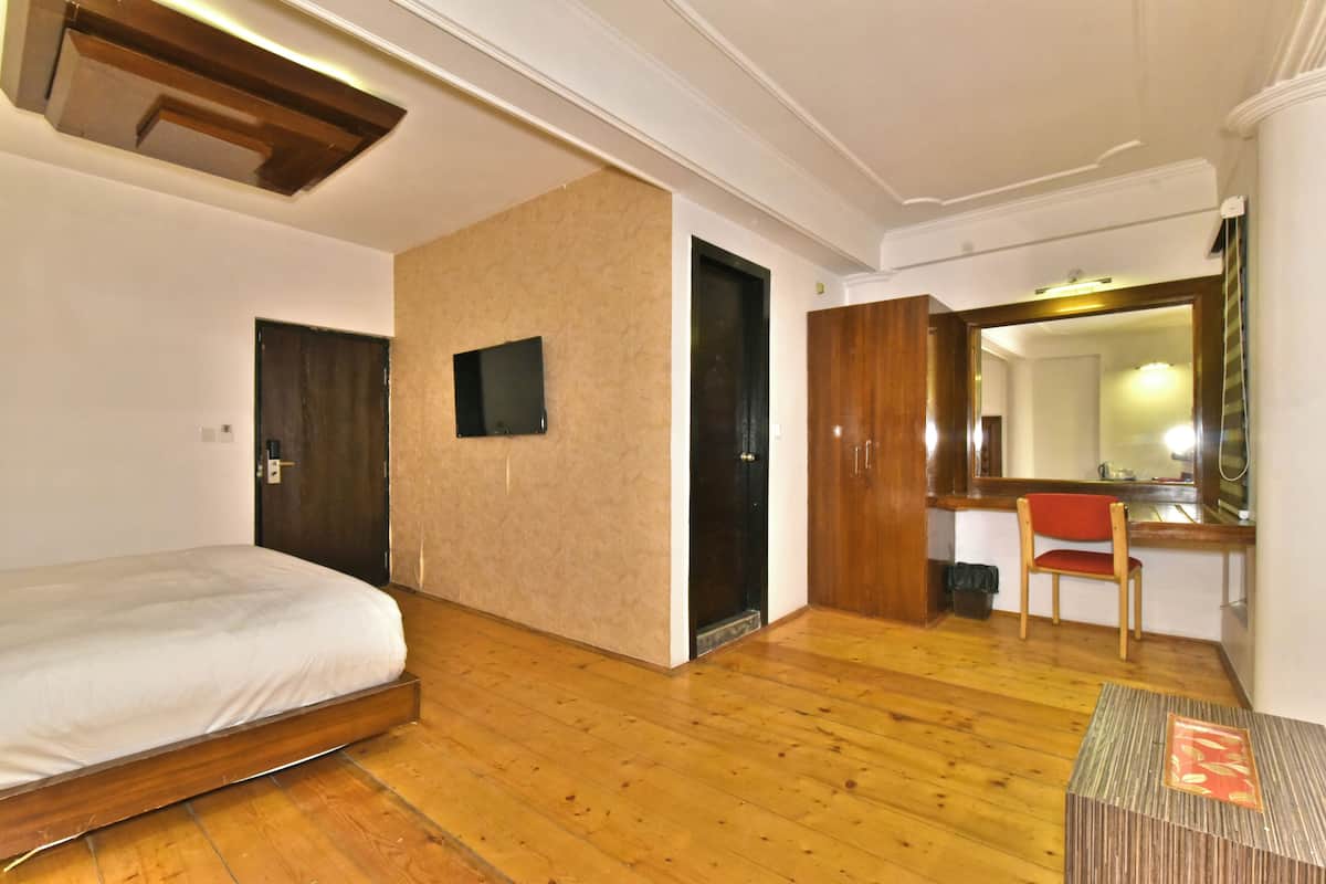 A hotel room with a wooden floor, a bed, a desk, a chair, a mirror, and a TV.