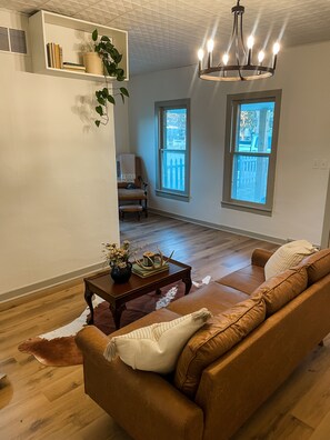 A living room with a brown leather sofa, a wooden coffee table with decorative items, a potted plant, and a chandelier.