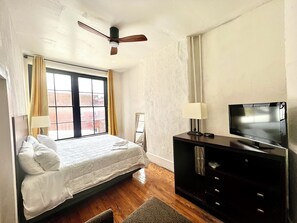 A hotel room with a bed, a television on a stand, a ceiling fan, and a window with curtains.