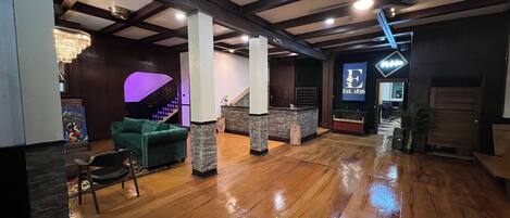 A spacious lobby with wooden flooring, a green sofa, a reception desk, and a staircase with purple lighting.