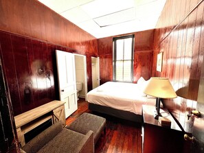 A bedroom with a wooden floor, a bed with a white comforter, a bedside lamp, a wooden bench, a window, and a door leading to a bathroom.