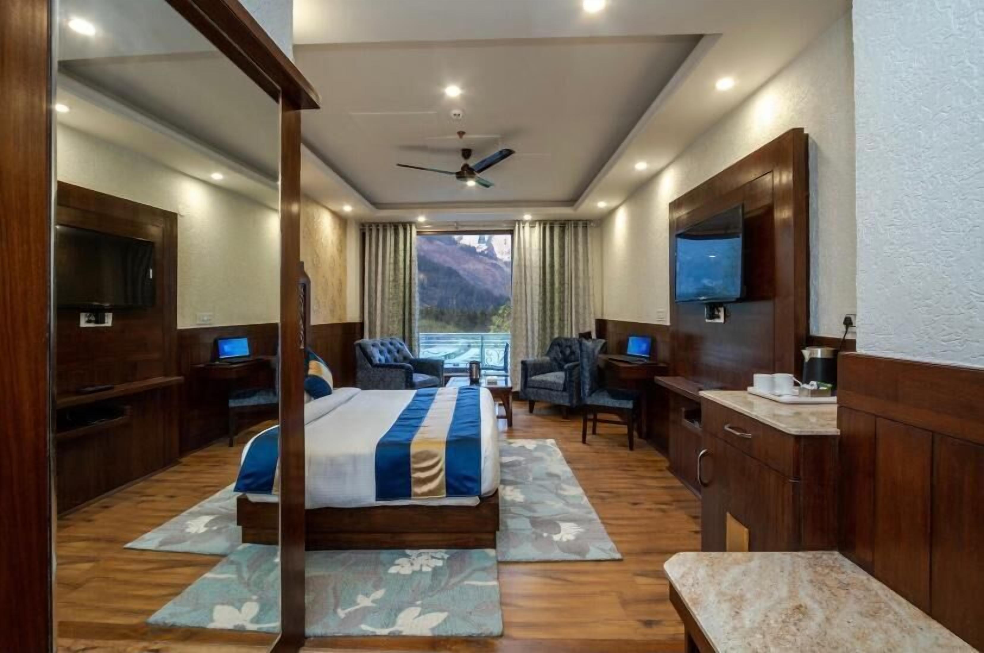 A hotel room with a bed, two armchairs, a desk, a TV, and a view of mountains.