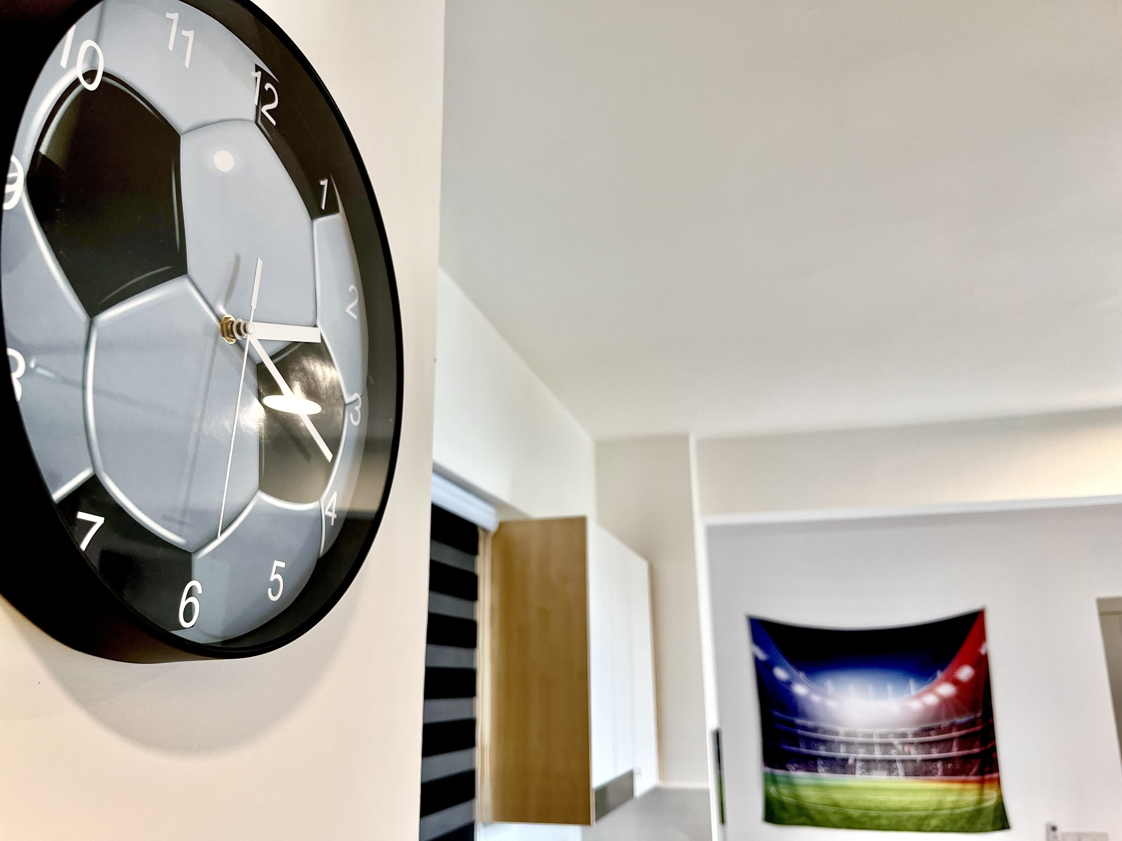 Eine Wanduhr mit sechseckigem Zifferblatt und den Ziffern 1 bis 12, eine Tür und ein Gemälde, das ein Sportstadion zeigt.