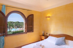 Une chambre d’hôtel comprenant un lit, une table de chevet, une fenêtre donnant sur une piscine et un espace verdoyant, ainsi qu’une lampe fixée au mur.