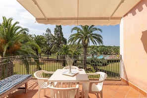 A balcony with a table, chairs, and a bench, overlooking a pool and palm trees.