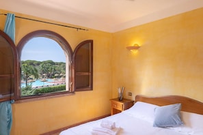 A hotel room with a bed, a nightstand, a window with a view of a pool and greenery, and a wall-mounted light. - Classical Residence Baia delle Palme Premium Bilo 4 nm1213 (Santa Margherita di Pula)