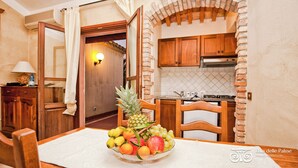A dining area with a table set for two, a fruit bowl, wooden chairs, a lamp, and a kitchen in the background.
