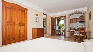 Een slaapkamer met een grote houten garderobe, een klein bureau met stoel, en uitzicht op de tuin door een open deur.