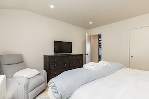 A bedroom with a bed, a grey sofa, a dark dresser, a television, and a door leading to a bathroom.