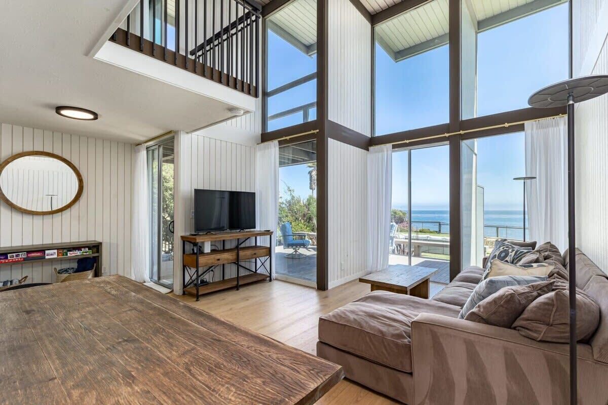 A spacious living area with a wooden table, a sofa, and a television. There is a balcony with a view of the sea.