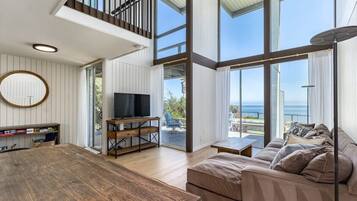 A spacious living area with a wooden table, a sofa, and a television. There is a balcony with a view of the sea.