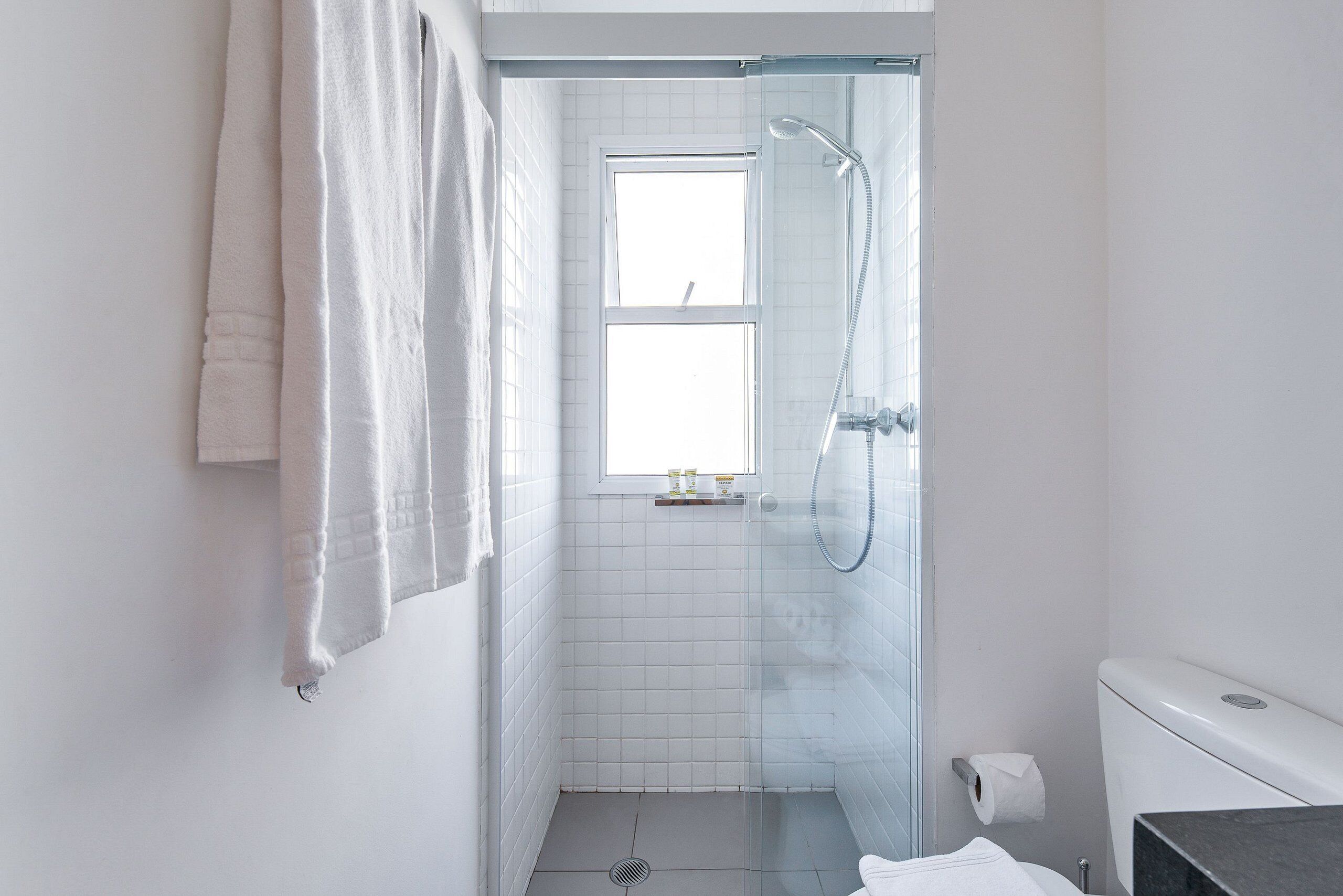 Une salle de bain avec une douche à parois de verre, des carreaux blancs, une fenêtre et des toilettes.