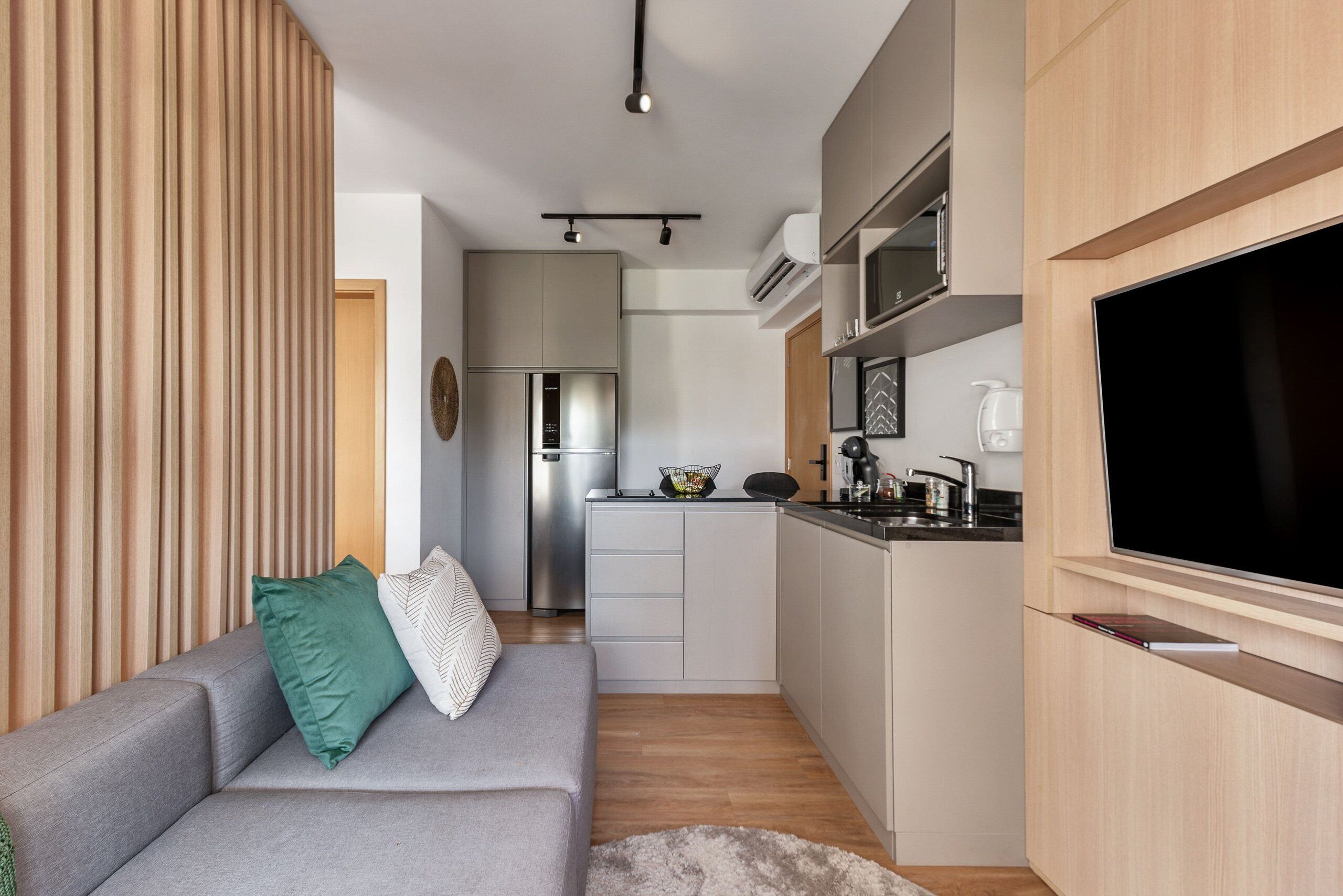 A compact living space with a kitchenette, a grey sofa with green and white pillows, and a flat-screen TV mounted on a wooden cabinet.
