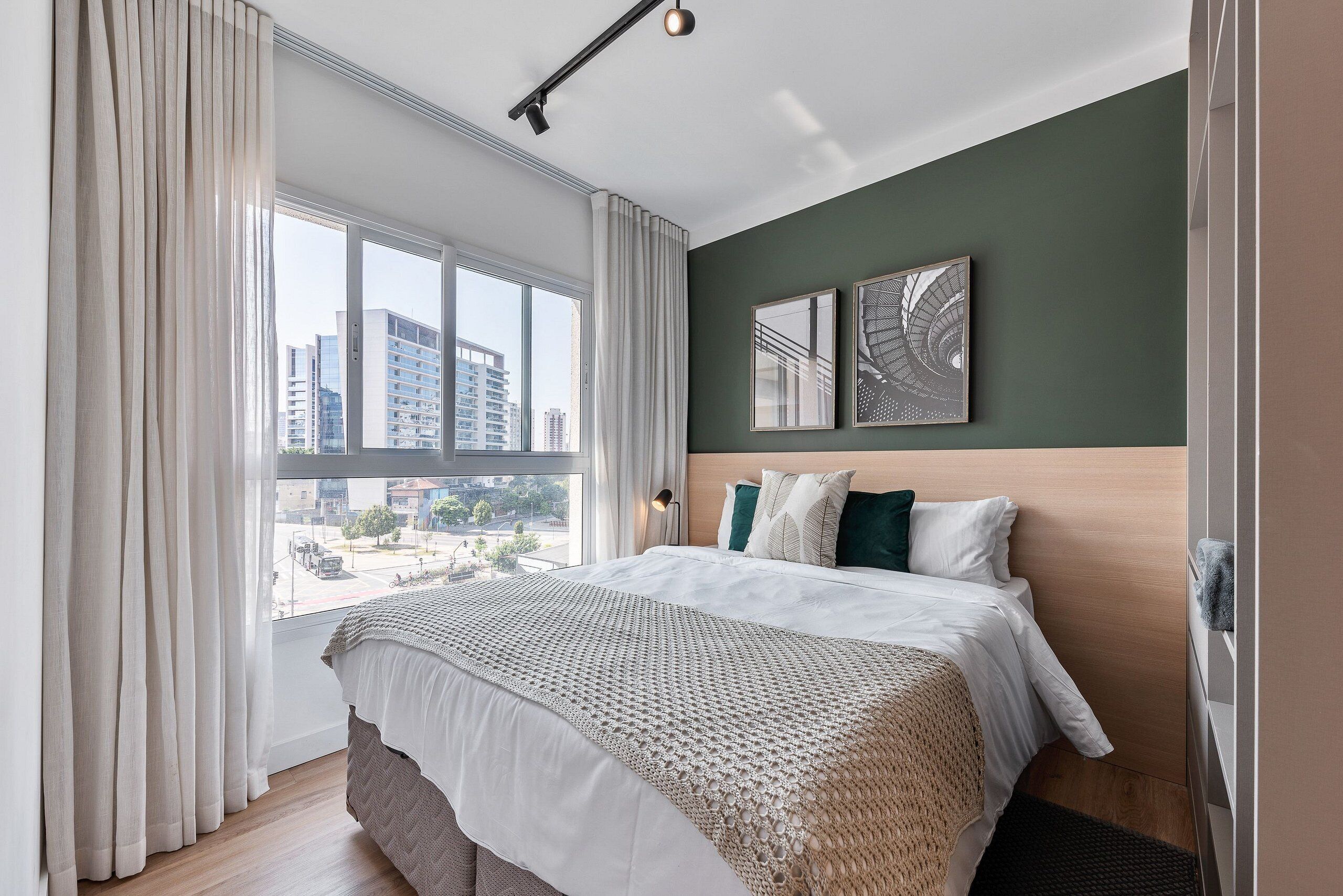 A modern hotel room with a large window, a bed with white linens, a wooden headboard, and two framed artworks on the wall.