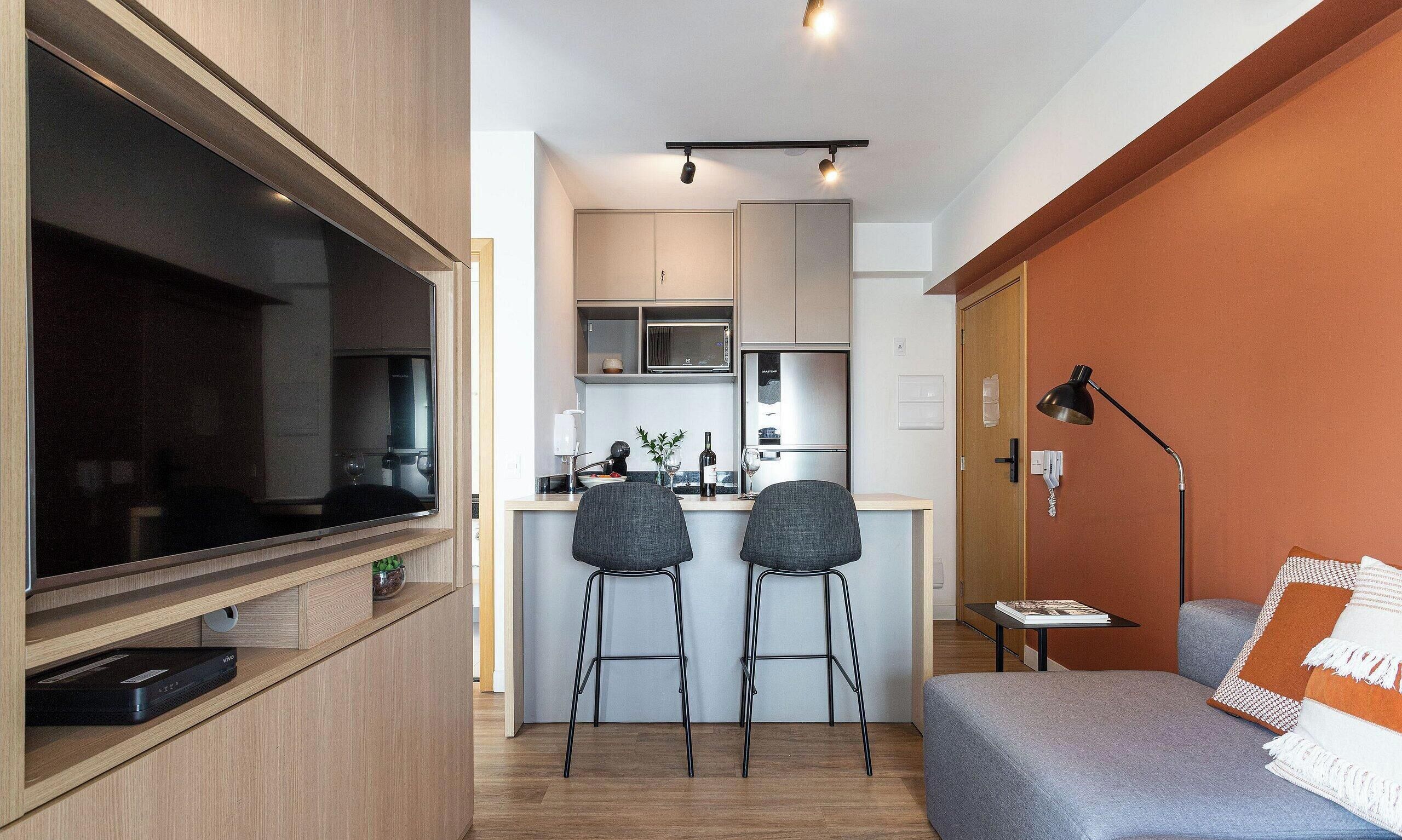 Un espacio habitable compacto con kitchenette, un televisor de pantalla plana, dos taburetes, un sofá y una lámpara.