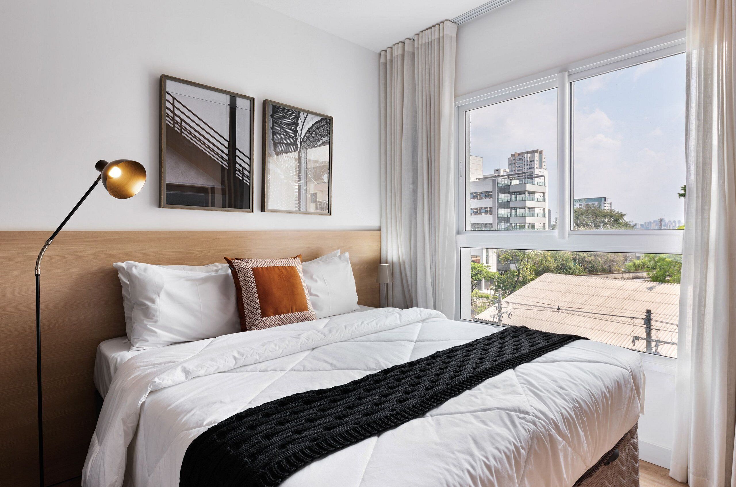 Habitación de hotel con una cama grande, una lámpara de pie, dos cuadros enmarcados y vistas de edificios a través de la ventana.