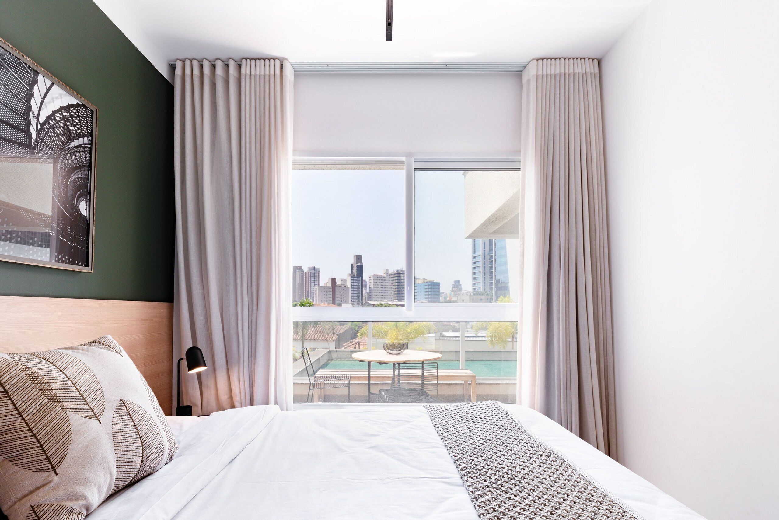 A hotel room with a bed, a desk, a chair, a lamp, and a view of a cityscape through large windows.
