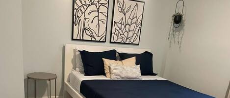 Un dormitorio con una cama, una mesita auxiliar, una silla y dos cuadros en la pared.
