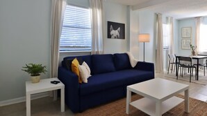 Een woonkamer met een blauwe bank, een witte salontafel, een bijzettafeltje met een plant, en een eethoek met een tafel en stoelen.