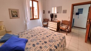 Una camera da letto con un letto, una cassettiera, due sedie, una finestra con tende, una ventola a soffitto e una porta che dà su un'altra stanza.